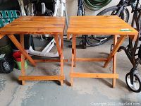Pair of matching folding wooden side tables positioned side by side on a concrete floor, showing overall condition and finish.