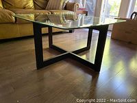 Angle view showing clear square glass top with beveled edges and rounded corners resting on black intersecting wood stand base on hardwood floor.
