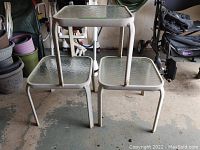 Three matching square glass top patio side tables with white metal frames shown stacked and arranged on a concrete floor.