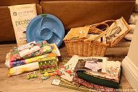 Full view of picnic basket with utensils, large blue plastic divided platter, folded tablecloths and placemats, storage bag, decorative items and tablecloth weights arranged on a couch.
