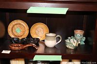 Photo of shelf displaying two round wooden decorative plates, pottery mug, ceramic spoon rest, set of five small black bowls, and additional pottery items including a greenish-blue bowl and a small pot figurine.