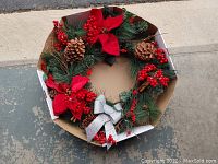 Full view of the 24-inch diameter wreath in cardboard box showing green pine needles, pinecones, red berries, red poinsettias and silver bow.