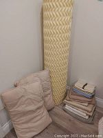 Full view of light yellow textured foam mattress topper standing upright with two light-colored quilted pillows and folded assorted bed sheets