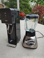 Photo showing Sodastream sparkling water maker and Oster blender side by side on an outdoor surface with garden background.