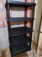 Large black plastic utility storage shelf with five perforated tiers, shown assembled in a storage area with unfinished walls and insulation visible behind.