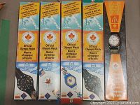 Five original McDonalds Olympic watch boxes in a row with one watch displayed outside its box, showing different designs and Olympic branding.
