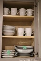 White mugs, shallow white bowls, and smaller white plates stacked in a cabinet shelf.