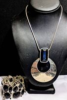 Photo of a large pendant necklace with a silver-tone crescent-shaped pendant and rectangular black rhinestone on a chain, displayed on a black bust.
