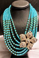 Full front view of the necklace displayed on a black mannequin highlighting multiple turquoise bead strands with gold-tone beads and a large gold-tone flower pendant with rhinestones and pearl accents.