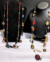 Three handblown glass bead necklaces with colorful chunky beads in different shapes, displayed on black and white stands (not included) against a black background.