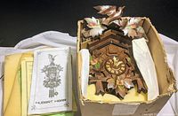 Hubert Herr Black Forest cuckoo clock in original box with visible instruction manuals beside it. Clock features carved wood with painted flowers and bird.