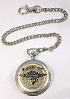 Front of the pocket watch displaying Harley Davidson logo with textured background and gold accents with chain attached.
