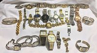 Full lot of 29 metal band watches arranged on a white surface showing watches with primarily silver and gold tones, many with linked and expandable bands.