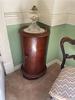 Cylindrical mahogany commode cabinet with marble top and glass lamp on top, positioned in a corner of a room