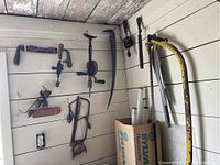 Wide view of various antique hand tools hanging on white panel wall including saws, hand drills, and a bow saw.