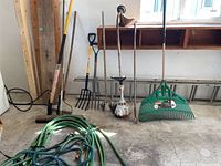 Photo of garden tools including hoses, rakes, pitchforks, shovel, broom, and Stihl weed wacker positioned against a garage wall.
