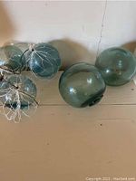 Five greenish-blue vintage glass fishing float balls, two wrapped with white nets, displayed on a wooden surface.