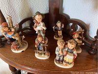 Wide view showing all six Hummel figurines arranged on a wooden chair seat, displaying size and condition.