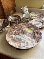 Photo showing stacks of large dinner plates, small side plates, bowls, and ceramic mugs with brown and cream Old English countryside patterns.