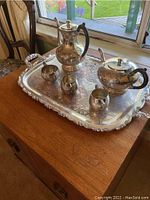 Silver plated tea and coffee service set arranged on a large ornate tray, showing the coffee pot, teapot, creamer, sugar bowl and a small cup with floral engraved detailing and black handles on the pots.