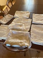 Six white or off-white folded tablecloths arranged on a wooden table, showing fabric texture and color.