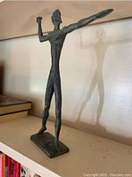 Front angled view of slender copper statue of Zeus in a stylized form, showing extended arm and rectangular base.