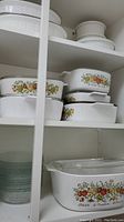 Multiple white ceramic baking dishes stacked on shelves with distinct vegetable pattern designs on sides, including artichokes, onions, and tomatoes.