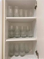 Three shelves of clear drinking glasses in a cabinet, various sizes and styles visible, some stacking, exhibiting geometric cut glass patterns.