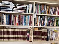 Full bookshelf view with the World Book Encyclopedia set volumes 1-12 on the bottom left and right shelves with the World Atlas in between. Adjacent shelves hold additional reference books and general non-fiction titles.