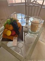 Photo showing faux fruit arrangement with oranges, lemon, green and black grapes, apple in wooden tray and adjacent glass candle holder with candle on glass table.