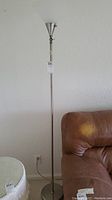 Full height view of the floor lamp standing beside sofa, showing slim silver metal pole and conical white plastic shade.