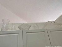 Several glass pieces and a plastic covered tray arranged on top of kitchen cabinets, including tall vases and a fan-shaped glass bowl.