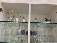Glassware including silver rimmed glasses, goblets, other assorted glasses, some with blue designs, displayed on glass shelves inside a cabinet.