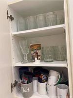 Three shelves of assorted glasses and mugs inside a white cabinet. Top shelf has tall clear ribbed glasses and short textured ones. Middle shelf has a mix of goblet style glasses, a tall printed glass, and short clear glasses. Bottom shelf has a variety of coffee mugs including black, white, and colorful mugs with prints and text.