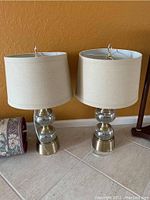Pair of glass and metal base table lamps with beige fabric shades shown side by side on floor against orange wall. One base has a visible line crack.
