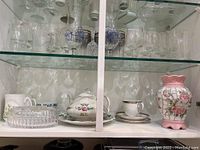 Glass cabinet shelf showing ceramic mosaic floral vase, ironstone teapot with floral designs, porcelain cups, and glass covered candy dish.