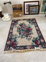 Full view of rectangular floral area rug showing beige background, burgundy and aqua floral pattern, dark floral border, and fringe edges, laid on tiled floor with some other household items around.