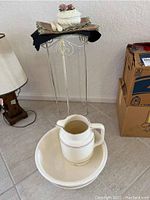 Round white ceramic pitcher sitting inside a large round ceramic bowl, both with gold rim detailing, placed on tiled floor. Metal table with white keepsake box on top in background.