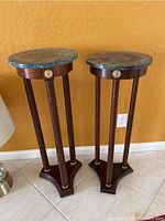 Front view showing two pedestal tables side by side against yellow wall, green marble tops and dark wooden bases with three vertical supports and gold medallions visible.