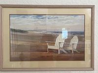 Framed wall art featuring a beach scene with two white Adirondack chairs looking out at the ocean. Light wood frame with cream matting under glass.