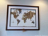 Framed world map wall art showing front view with centered continental map in brown hues on white background, within a brown wooden frame and white matting.