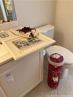 View showing three framed vintage black and white wall art pieces with cream-colored frames resting on bathroom counter, toilet, and a burgundy fabric-covered toilet paper holder next to the toilet with white lace and rose decorations.