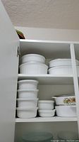 Photo showing upper shelves with multiple white ceramic CorningWare baking dishes and casseroles including round ribbed sides and oval casserole with floral design
