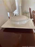 Side angle view of white ceramic tureen with lid on top of large rectangular serving platter on wooden surface.