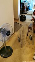 Floor fan and metal folding cart positioned side by side on carpeted floor indoors, showing both items in full.