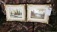 Two framed landscape paintings resting on patterned cushions showing both artworks and their golden carved frames.