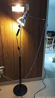 Black floor lamp lit with two adjustable light heads and round base on carpeted floor in front of wood panel wall.