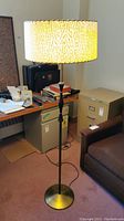 Full view of mid-century modern floor lamp with lit textured fiberglass drum shade casting warm yellow light. Lamp standing near filing cabinets and sofa.
