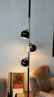 Full view of mid-century modern black tension lamp with three spherical lights mounted vertically on a pole between two chairs. Lamp is turned on.