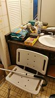 Wide view showing bathroom countertop with multiple personal care items, unopened disposable underpads, and white plastic shower seat with backrest in front of sink.